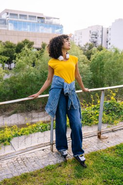 Kıvırcık saçlı, kulaklık takan genç bir kadın şehir parkında yürüyor, müziğin ve temiz havanın tadını çıkarıyor.