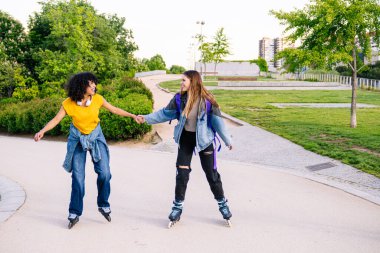 İki bayan arkadaş şehir parkında el ele tutuşarak ve gülümseyerek paten kaymaktan zevk alıyorlar.