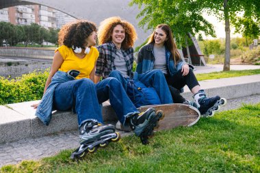 Üç genç patenci arkadaş birlikte neşeli bir parkta gülüyor ve kaygısızca vakit geçiriyorlar, güneşli bir günü ve güzel havayı içlerine çekiyorlar.