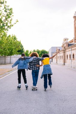 Birbirine sarılmış bir grup arkadaş sokakta paten ve kaykayla yürüyorlar, öğleden sonranın tadını çıkarıyorlar.