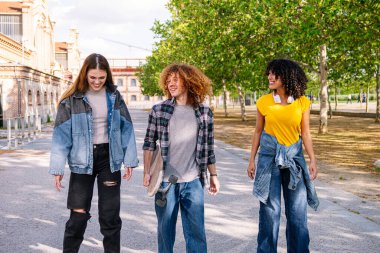 Üç mutlu arkadaş parkta yürüyorlar, sohbet ediyorlar ve güneşli bir öğleden sonra bir kaykay taşıyorlar.