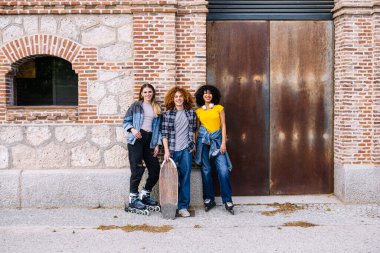 Üç arkadaş birlikte bir öğleden sonranın tadını çıkarıyor, kaykay ve paten ile şehir ortamında tuğla bir duvara ve paslı metal bir kapıya karşı poz veriyorlar.
