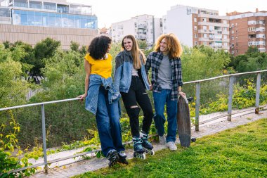 Üç arkadaş şehir parkında eğleniyor, biri kaykaylı, diğeri patenli, öğleden sonranın keyfini çıkarıyorlar.