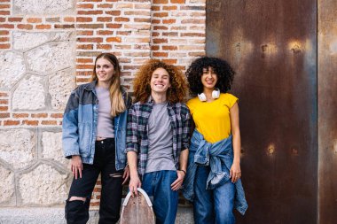 Üç neşeli genç öğrenci duvara karşı poz veriyor, biri kaykay tutuyor, birlikte boş zamanlarının tadını çıkarıyorlar.