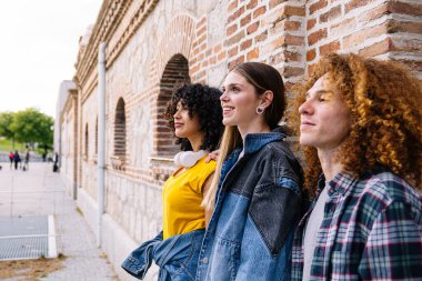 Üç genç arkadaş, muhtemelen patenciler, bir tuğla duvarın yanında dururlar, öğleden sonranın tadını çıkarırlar ve kaykay parkına doğru bakarlar.