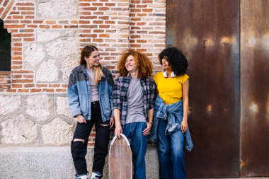 Üç arkadaş açık havada kaykay yaparken keyifli bir anı paylaşıyor, şehir duvarına yaslanıyor, dostluk ve macera hissi yayıyor.