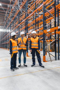 Güvenlik yelekleri ve şapka giyen depo çalışanları büyük bir dağıtım merkezinde lojistik ve envanter yönetimini tartışıyorlar.