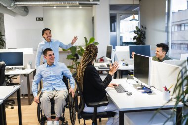 Çeşitli meslektaşlar modern bir çalışma alanında birbirlerini selamlıyor, engelli bir iş arkadaşıyla birlikte tekerlekli sandalyede takım çalışmasını ve dahil edilmesini kutluyorlar.