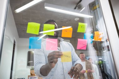 Gülümseyen yönetici cam duvara renkli yapışkan notlar yapıştırıyor, modern bir ofiste ortak bir proje toplantısında fikirleri paylaşıyor.