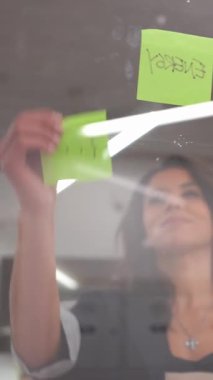 Genç iş kadını iki yapışkan nota eko ve enerjiyi ağır çekimde cam duvara yapıştırıyor.