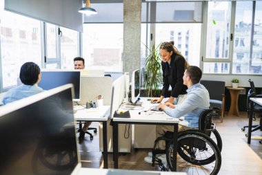 Çeşitli takımlar, parlak, modern bir ofiste birlikte çalışıyorlar. İşbirliği, dahil etme ve asist teknolojisi sergiliyorlar. Tekerlekli sandalye kullanıyorlar.