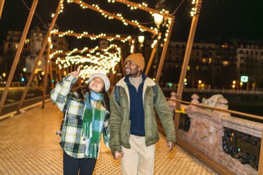 Genç, çok ırklı bir çift, kış tatili boyunca köprüde el ele yürürken Noel ışıklarına hayranlık duyuyor.