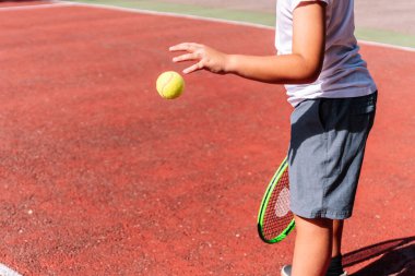 Çocuk kırmızı kilden bir kortta tenis topu servis etmeye hazırlanıyor, raketi tutuyor ve topu havaya fırlatıyor.