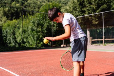 Yoğunlaşmış genç bir çocuk kırmızı kortta tenis oynamaya hazırlanıyor. Gençlik sporları ve yarışma ruhunu somutlaştırıyor.