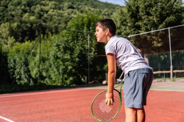 Tenis raketi ve top tutan konsantre çocuk yeşil ağaçlarla çevrili kırmızı bir kil kortta hizmet vermeye hazır.