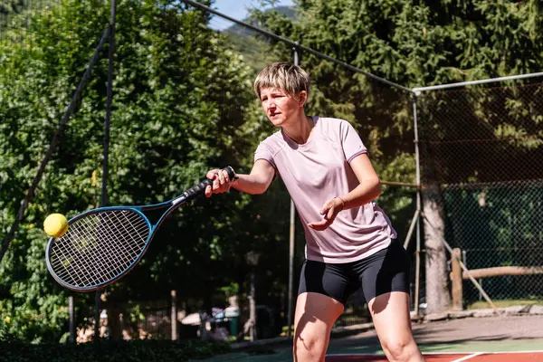 Konsantre kadın tenisçi ağaçlarla çevrili güneşli bir sahada forehand vuruyor.
