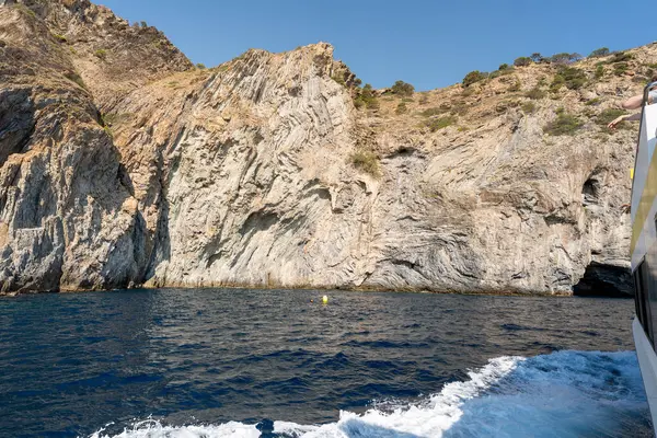 Güneşli bir yaz gününde kayalık güllerin kıyılarında yol alan motorlu tekne beyaz bir su izi bırakıyor.