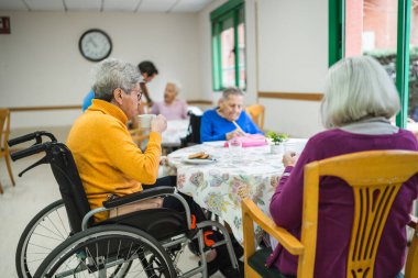 Huzurevindeki bir masada oturan yaşlılar, bazıları kahve içiyor, bazıları resim çiziyor, sosyal hayatın tadını çıkarıyor.