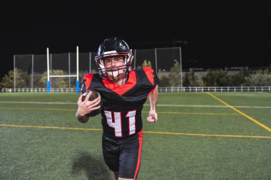 Gece maçında kaskla koşan üniformalı Amerikan futbolcu yeşil bir sahada topu taşıyor.