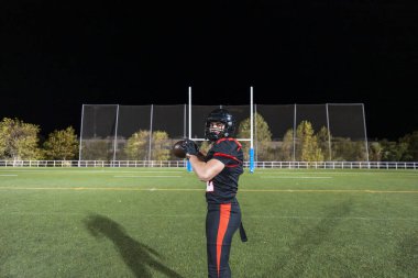 Erkek Amerikan futbolcusu üniformalı, kasklı ve top tutan yeşil bir sahada gece kale direkleri olan.