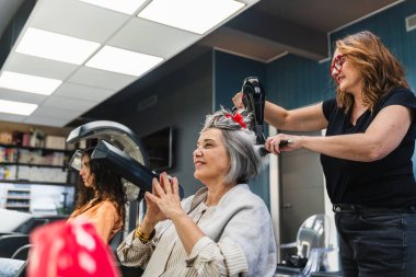 Kuaför modern bir güzellik salonunda yaşlı bir kadının gri saçlarını kurutuyor ve şekillendiriyor. Mutlu bir güzellik anı yaratıyor.