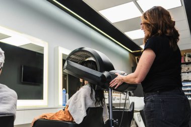 Saç bakım ve güzellik hizmetlerine odaklanan kukuletalı bir kurutma makinesinin altında saçını kurutan bir kadına yardım eden kuaför.