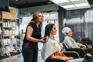 Kuaför genç bir kadına kuaförlük yapıyor. Bir diğer müşteri de arka planda kuaförde bekliyor.