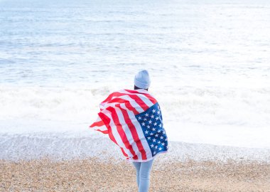 Mavi kazaklı ve şapkalı esmer kadın. Deniz kıyısında Amerikan bayrağı taşıyor.