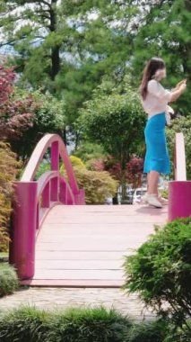 Mavi elbiseli genç esmer kadın akıllı telefonuyla fotoğraf ve video çekiyor. Pembe köprüdeki parkta. 