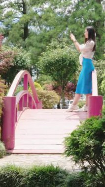 Mavi elbiseli genç esmer kadın akıllı telefonuyla fotoğraf ve video çekiyor. Pembe köprüdeki parkta. 