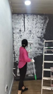 Pembe elbiseli esmer kadın bir tuğla duvarı boyuyor, siyahtan beyaza çeviriyor..