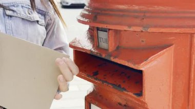 Şehir caddesinde mektupları klasik kırmızı posta kutusuna postalayan genç bir kadın.