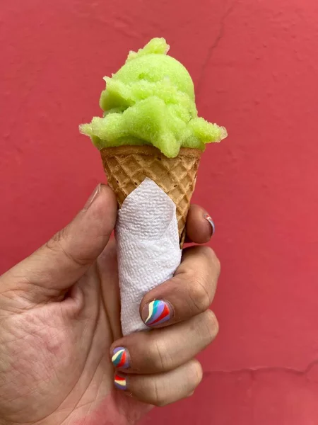 stock image Man's hand is holding a green, lime scoop of ice-cream in a cone against a red wall. Man has a nail-art manicure.