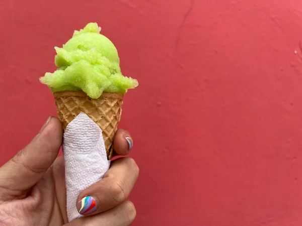 stock image Man's hand is holding a green, lime scoop of ice-cream in a cone against a red wall. Man has a nail-art manicure.