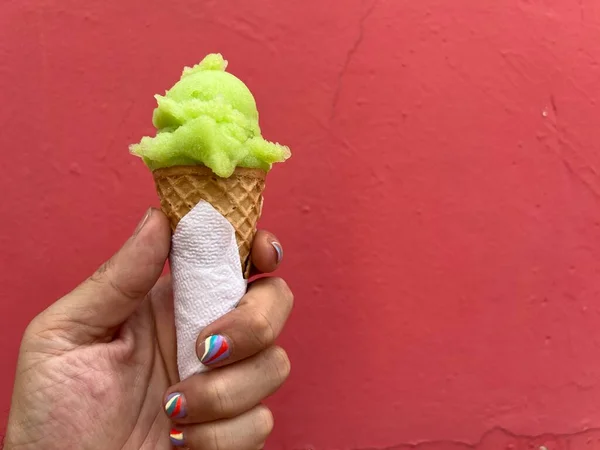 Man\'s hand is holding a green, lime scoop of ice-cream in a cone against a red wall. Man has a nail-art manicure.