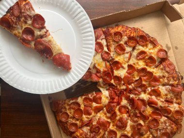 Karton bir kutuda peynirli, pepperonili büyük bir pizzadan beyaz bir kağıt tabakta pizza dilimi. Bir restorandan paket servis..