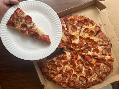 Karton bir kutuda peynirli, pepperonili büyük bir pizzadan beyaz bir kağıt tabakta pizza dilimi. Bir restorandan paket servis..