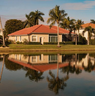 Güneşli bir günde Güney Florida 'da güzel bir yazlık. Florida 'nın güneybatısındaki bir gölün kıyısındaki tipik beton ev. Palmiye ağaçları, tropik bitkiler ve çiçekler, çimenler ve çam ağaçları. Florida.