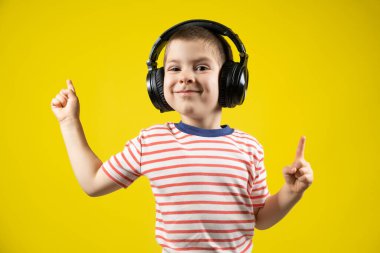 Kulaklıklı ve tişörtlü mutlu çocuk sarı bir arka planı işaret ediyor..