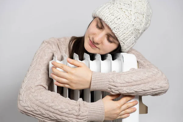 A brunette woman in a hat hugs an oil heater, warms up in winter