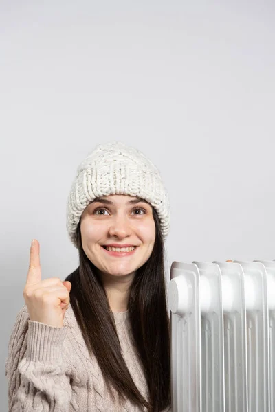 A woman in a hat getting warm near the oil heater and looks and points up with her finger
