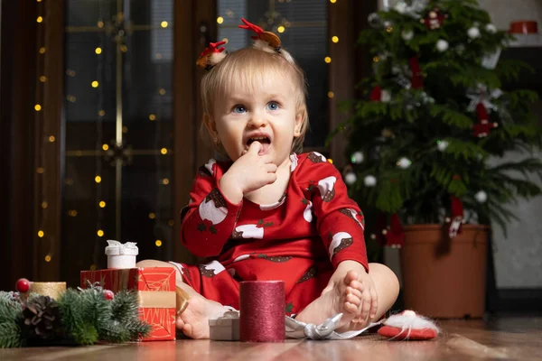 A one-year-old girl with blue eyes smiles sitting near a Christmas tree. Christmas, New Year and children