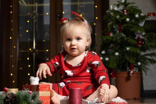 A one-year-old girl with blue eyes smiles sitting near a Christmas tree. Christmas, New Year and children