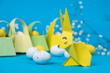 Kağıttan, origamiden ve mavi arka planda beyaz bıldırcın yumurtasından yapılmış bir Paskalya tavşanı heykeli..