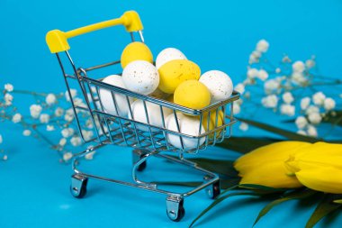 Easter white and yellow quail painted eggs in a shopping cart on a blue background. Easter Sale and shopping.