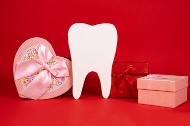 Valentine's Day in dentistry - a large white tooth, a heart and boxes of gifts on a red background, a place for text.