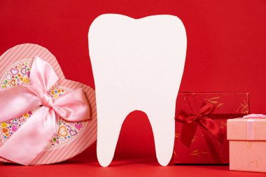 Valentine's Day in dentistry - a large white tooth, a heart and boxes of gifts on a red background, a place for text.
