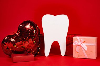 Valentine's Day in dentistry - a large white tooth, a heart and boxes of gifts on a red background, a place for text.