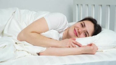 A woman lies in bed on her side on a white stripe satin bed linen and smiles quite a looking into the camera
