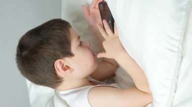 A 5 year old boy plays games on his phone while lying in bed. Gadgets in bed before bedtime in a child.